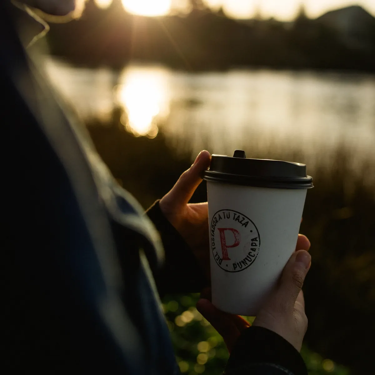 Mano sosteniendo un vaso de café Panaka frente a un lago al atardecer.