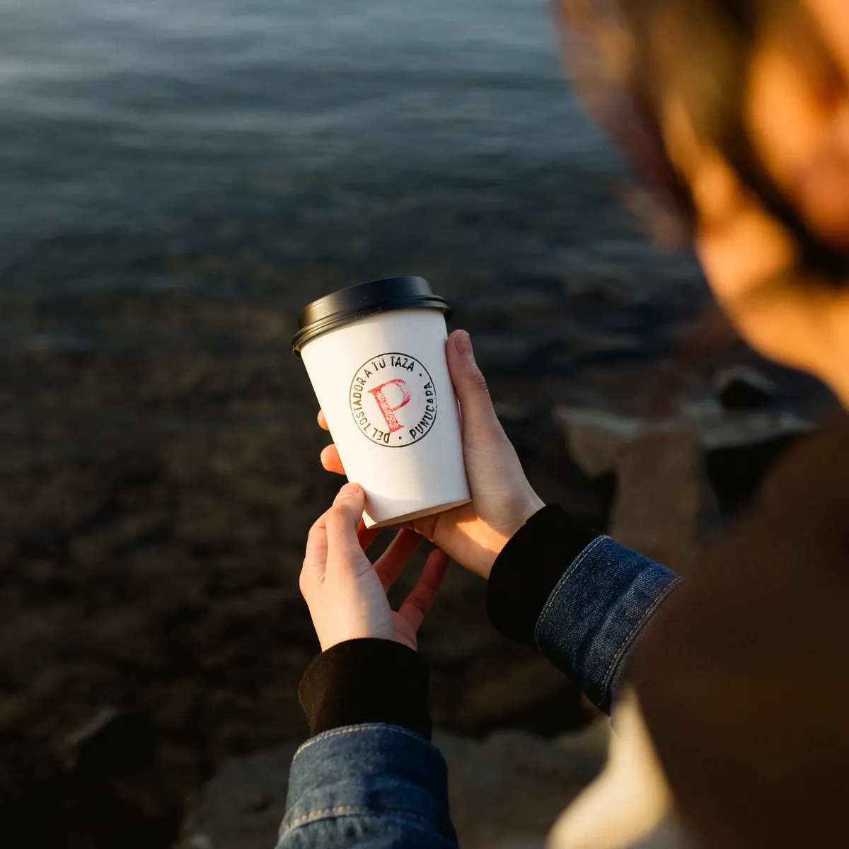 Mano sosteniendo un vaso de café Panaka junto al agua.