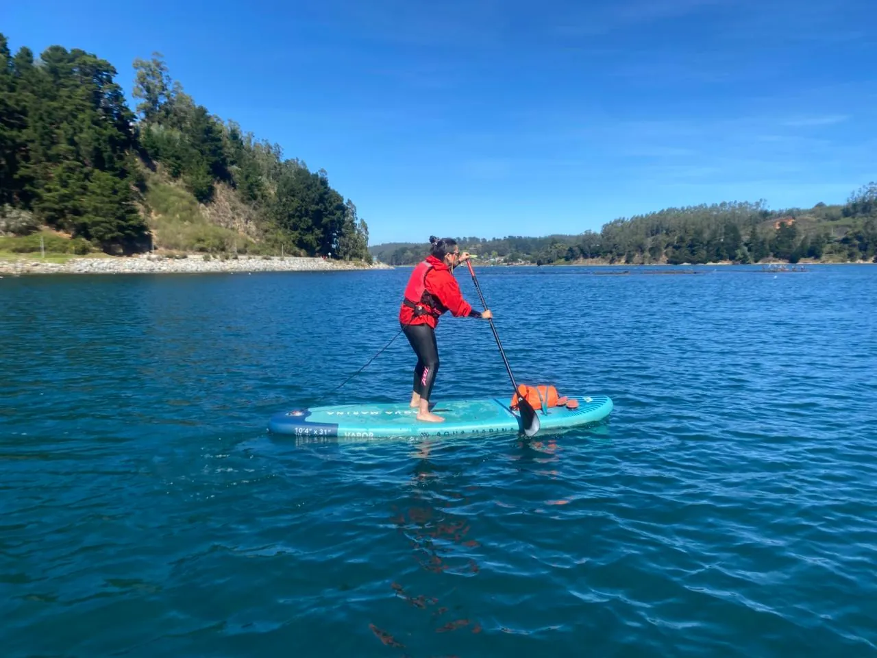 Isabel remando de pie en una tabla de SUP sobre agua azul, con chaqueta roja y chaleco salvavidas, entre cerros con bosque.